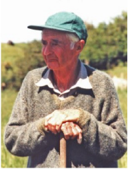 An elderly white man pictured outdoors, with a green field and trees in the background behind him. He is wearing a brown knitted jumper with a white collared shirt, and a green baseball cap. He is looking out of shot and resting his hands on the handle of a wooden tool in front of him.